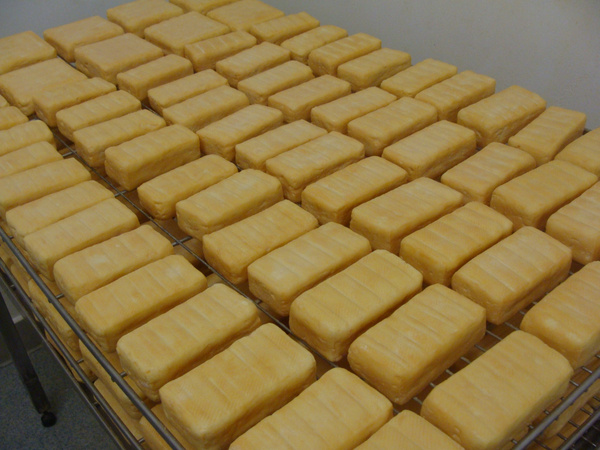 KI generiert: Reihen gelber rechteckiger Käseblöcke lagern auf Edelstahlregalen.