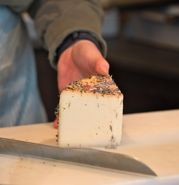 KI generiert: Eine Person hält ein Stück Käse mit Kräutern oben drauf. Ein Messer ist im Vordergrund.