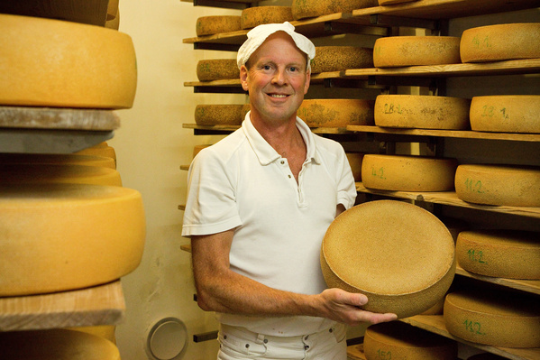 KI generiert: Ein Mann hält einen Käselaib in einem Lager mit vielen Käselaiben.